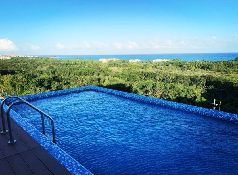 An inviting infinity pool extends towards the horizon, blending seamlessly with the lush green landscape and distant ocean views. Sunlight reflects off the water, creating a calm and serene environment for relaxation and enjoyment.
