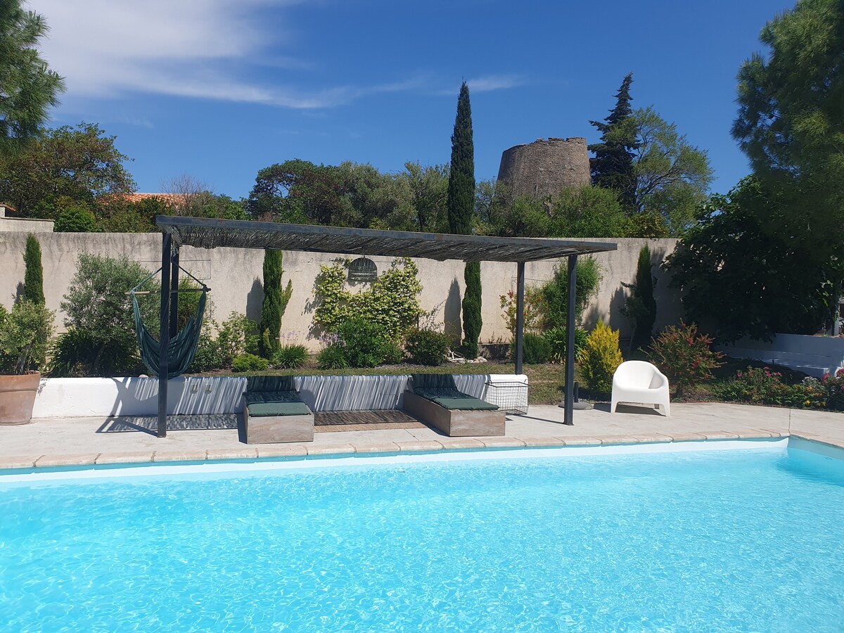 A clear swimming pool reflects the blue sky, surrounded by a sun patio featuring shaded lounging areas and greenery. A white chair sits nearby, and a historic stone structure is visible in the background, alongside neatly arranged plants and trees that provide privacy.