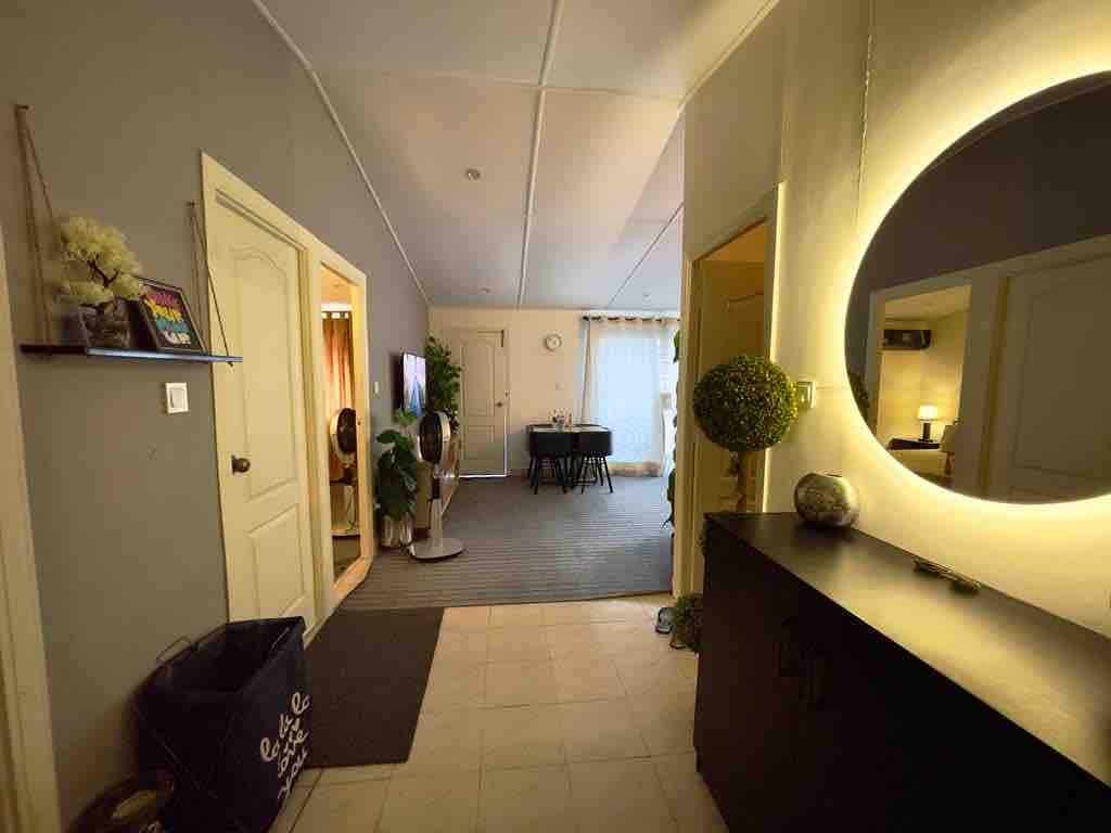 A welcoming entrance area is highlighted by a circular mirror and soft ambient lighting. A console table holds decorative items, and a fan is positioned nearby. The space leads to a dining area with a table set for four and a view of a window with sheer curtains.