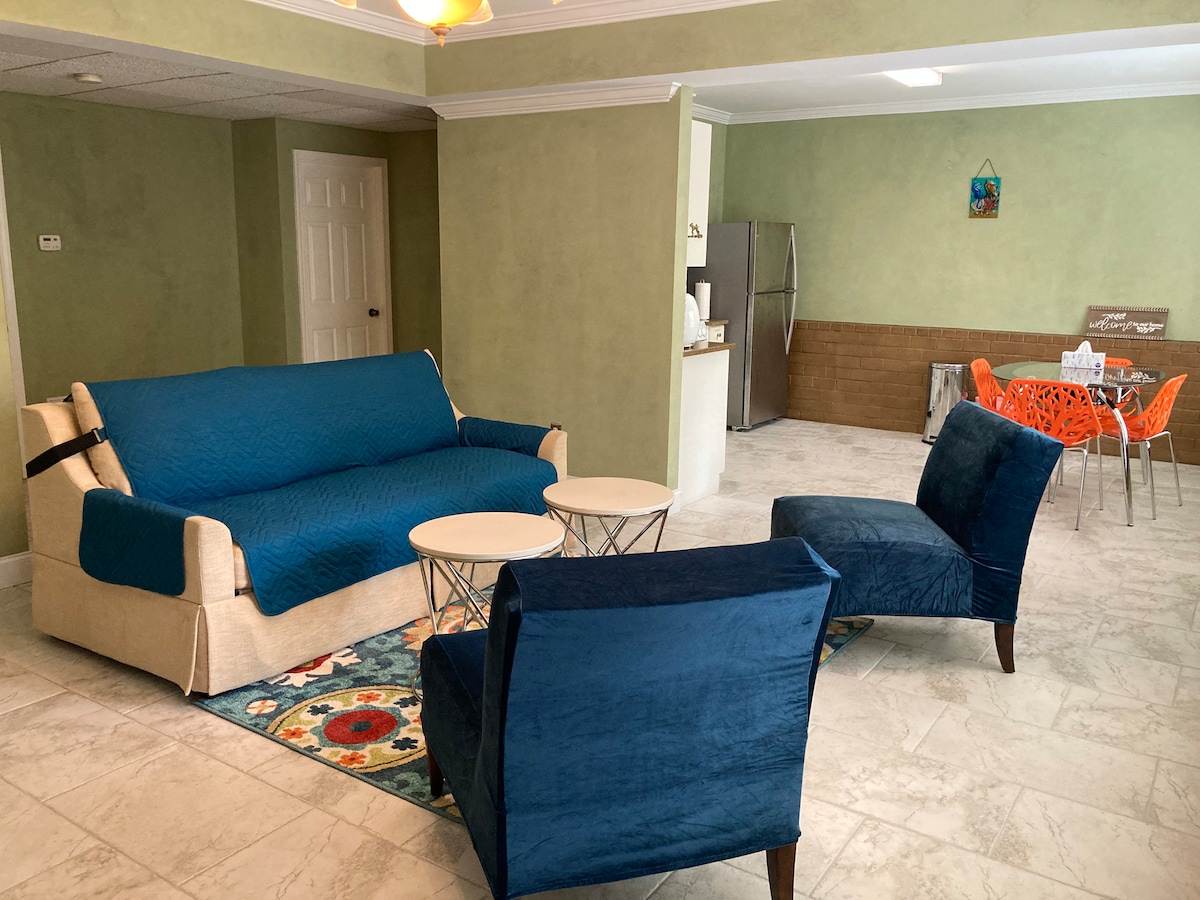 A spacious living area features a memory foam sofa-bed draped in a blue cover, accompanied by two plush chairs. A patterned area rug lies under a pair of round coffee tables. A dining table with colorful chairs is visible in the background, alongside a full kitchen.