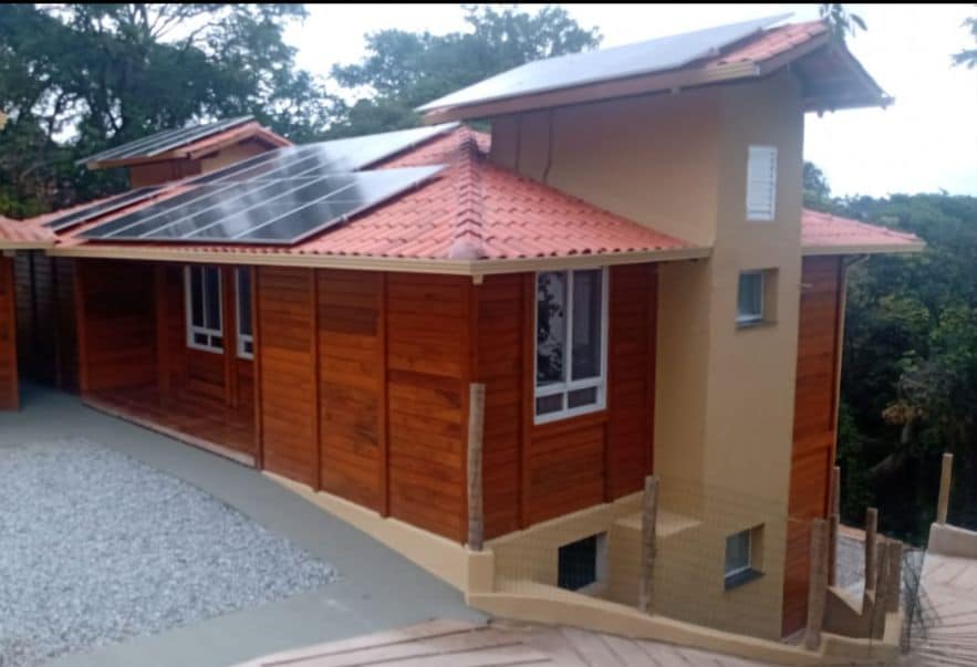 The exterior of a modern chalet is presented, featuring a combination of wooden paneling and a light-colored facade. Solar panels are installed on the sloped roof, and a gravel pathway leads to the structure. Large windows allow for natural light and views of the surrounding greenery.