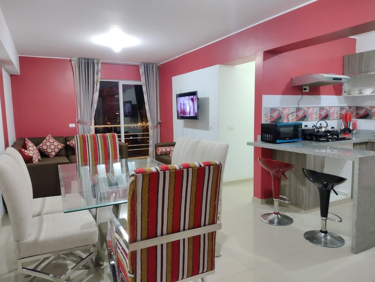 A contemporary open-concept living space features a glass dining table surrounded by striped chairs. A stylish kitchen with modern appliances is visible in the background. A comfortable seating area, adorned with colorful cushions, is enhanced by large windows that allow natural light to fill the room.