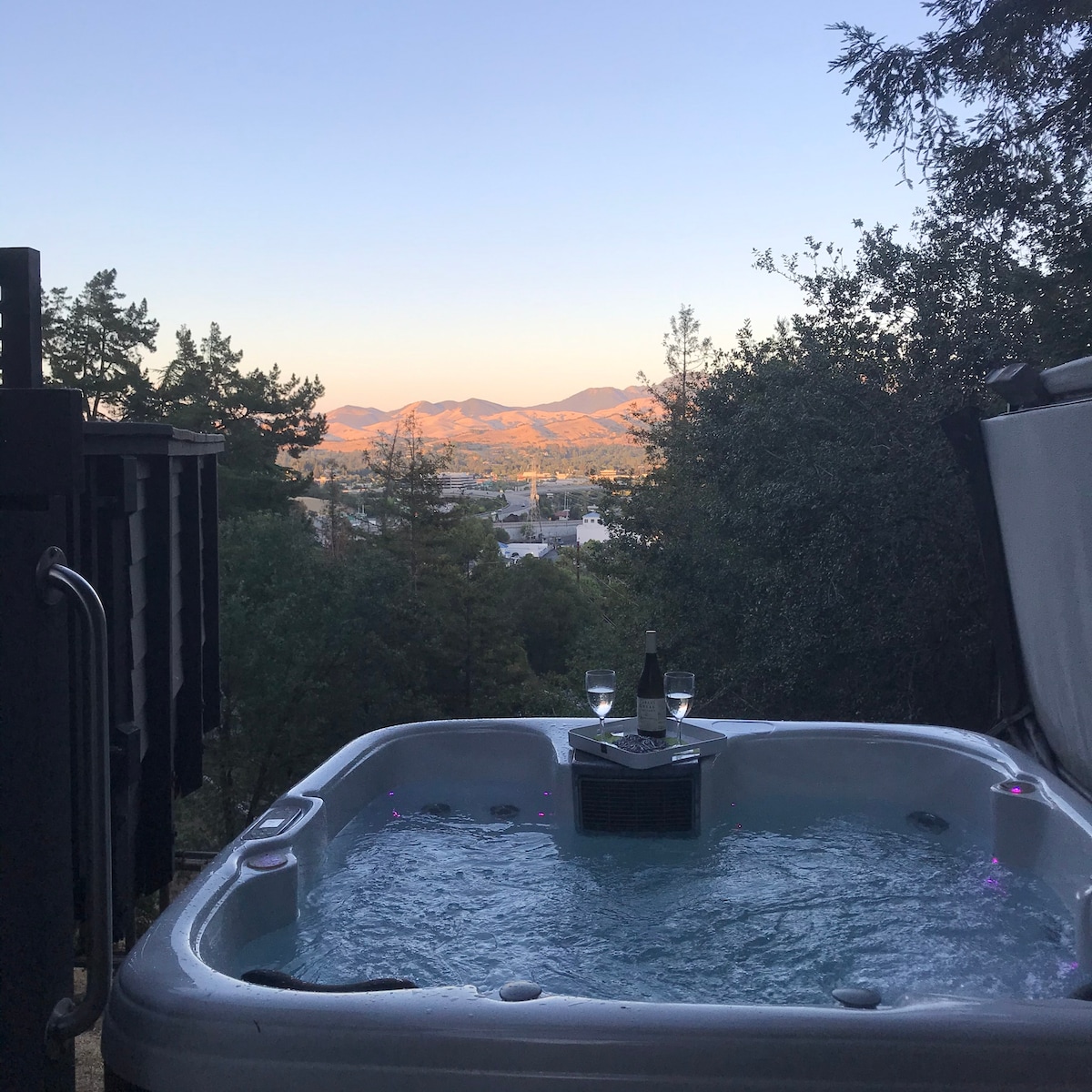 A private hot tub is situated in a shaded outdoor area, overlooking scenic mountain views. Two glasses and a bottle of wine are placed on the edge of the tub, while soft lighting enhances the serene atmosphere during dusk.