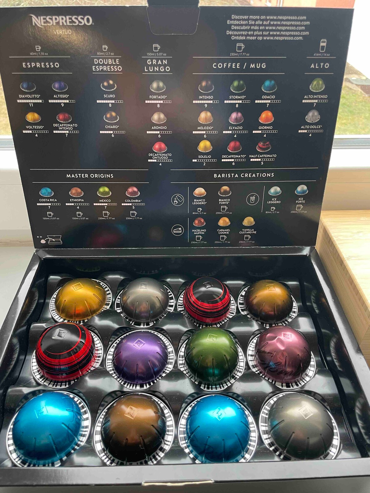 An organized display of Nespresso coffee capsules is presented in a neatly arranged box, featuring various colors and patterns. The capsules are arranged in trays, with labels indicating different types and flavors positioned above, showcasing a wide selection for coffee enthusiasts.