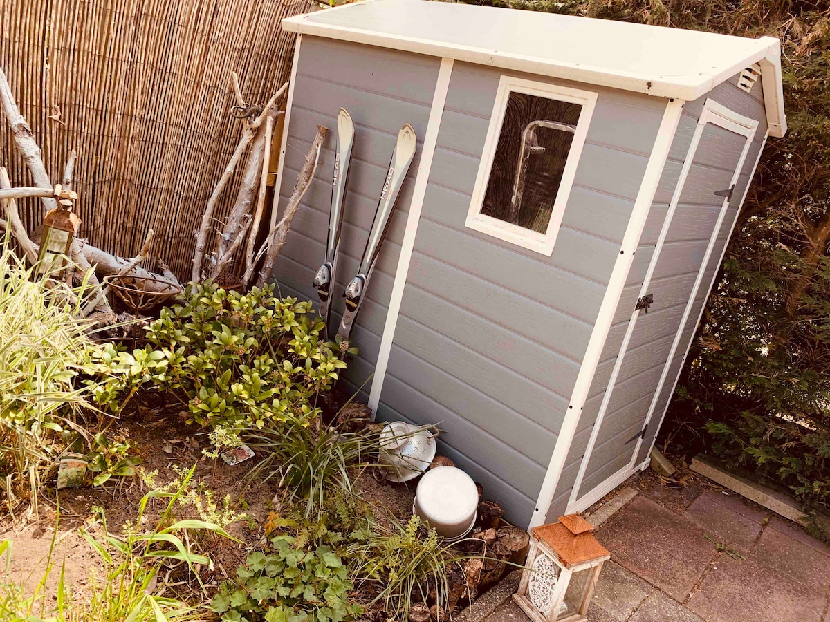 A small, slanted storage shed is nestled in a garden area, surrounded by low greenery and stone tiles. The shed features a single window and is positioned among various plants, including grasses and foliage. Nearby items include a lantern, a white bowl, and a few decorative elements.