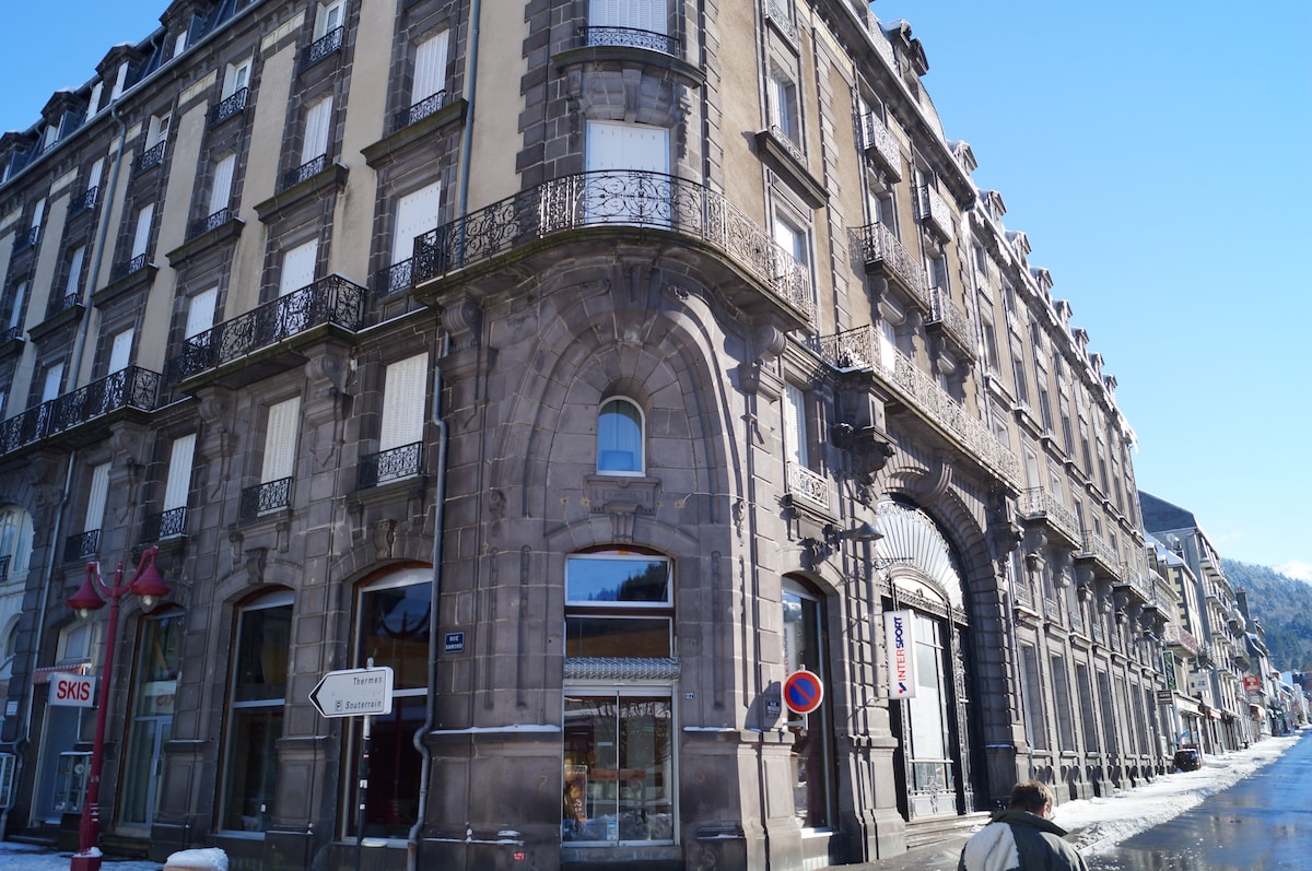 The historic building features ornate stone architecture with arched entrances and intricate wrought iron balconies. Large windows are present, and the structure showcases a blend of classical design elements. The surrounding area includes signage and a pathway, indicating its central location in Mont Dore.