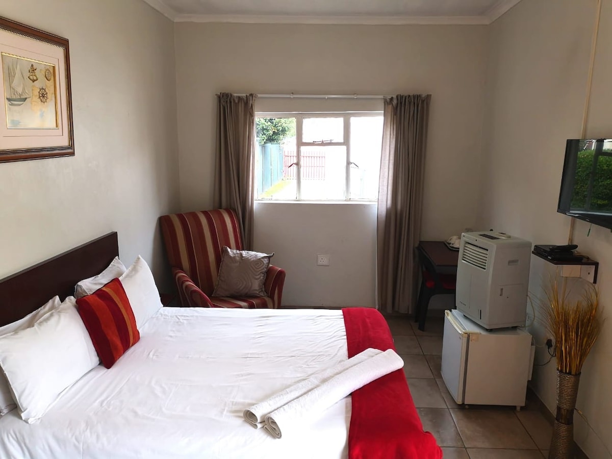 A cozy room contains a spacious bed dressed in white linens with red accent pillows. A striped armchair adds a touch of comfort near a window, which is dressed with light curtains. An air conditioning unit and a small desk are positioned against the wall.