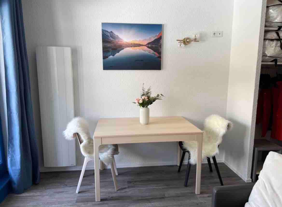 A small dining area features a light wooden table with two uniquely styled chairs. A simple vase of flowers sits at the center, while a scenic mountain landscape is displayed on the wall above. Natural light filters in through a nearby window, enhancing the inviting space.