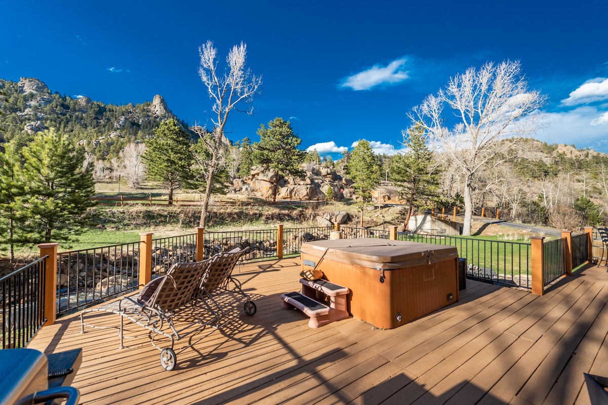 A spacious deck features a hot tub and lounge chairs, set against a backdrop of trees and rocky terrain. The clear blue sky enhances the outdoor space, inviting relaxation and enjoyment of the surrounding natural beauty.