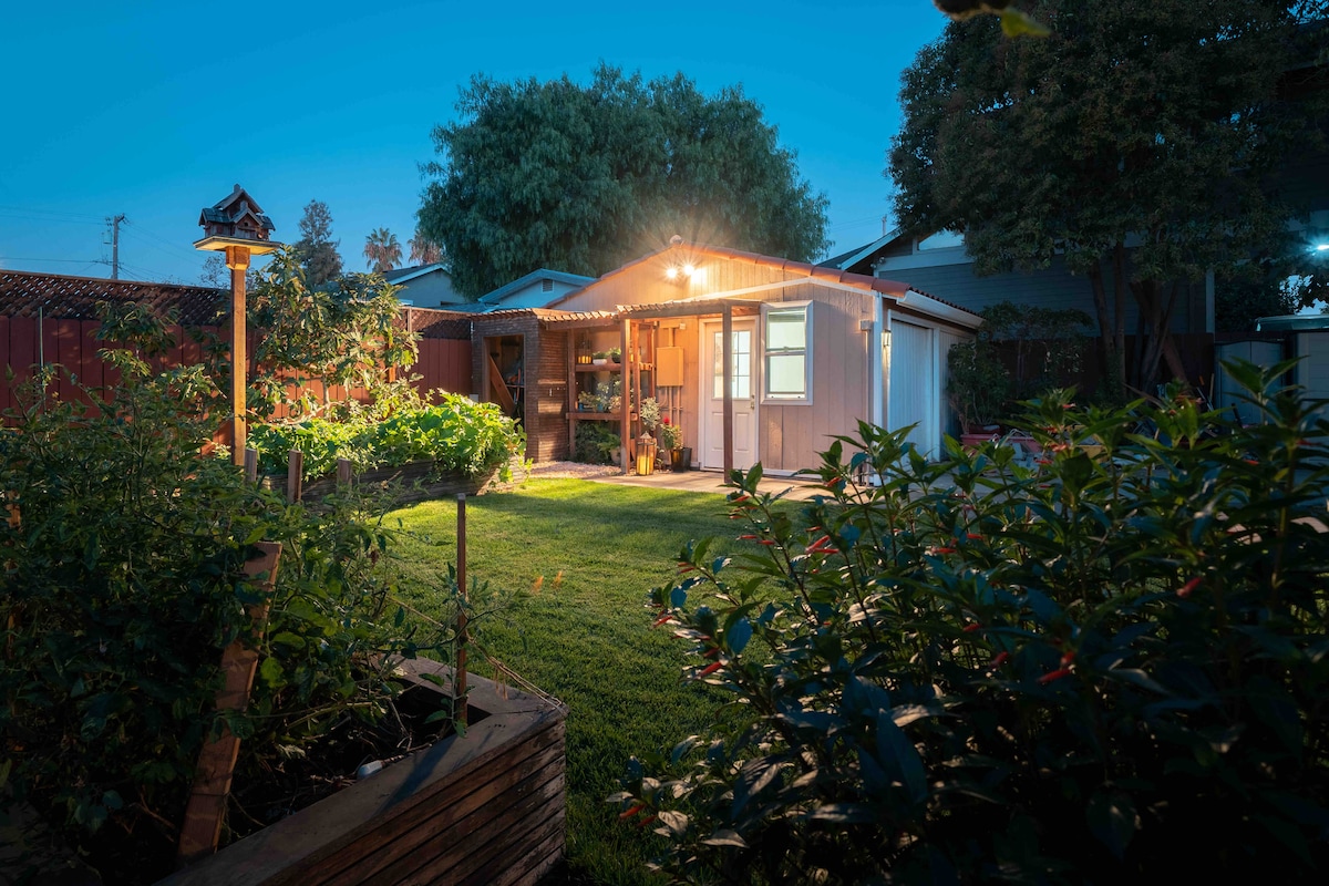A charming guest house is illuminated in the evening, surrounded by well-maintained greenery and garden beds. Soft light glows from the structure, highlighting a welcoming entrance and a peaceful outdoor space for relaxation.