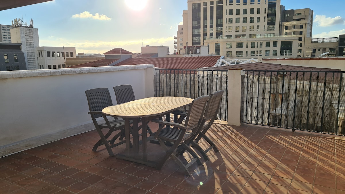 A private roof terrace is showcased with a round wooden table surrounded by four folding chairs. The sun casts a warm glow over the space, while a view of nearby buildings is visible in the background.