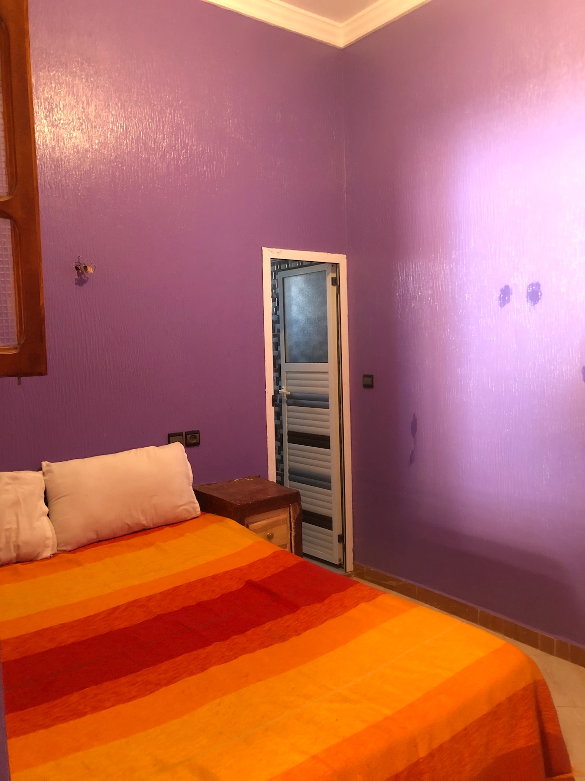 A bedroom is depicted with vibrant purple walls. A spacious bed is adorned with an orange and red striped blanket, complemented by soft white pillows. A door leads to an adjoining area, while natural light illuminates part of the room, enhancing its cozy ambiance.