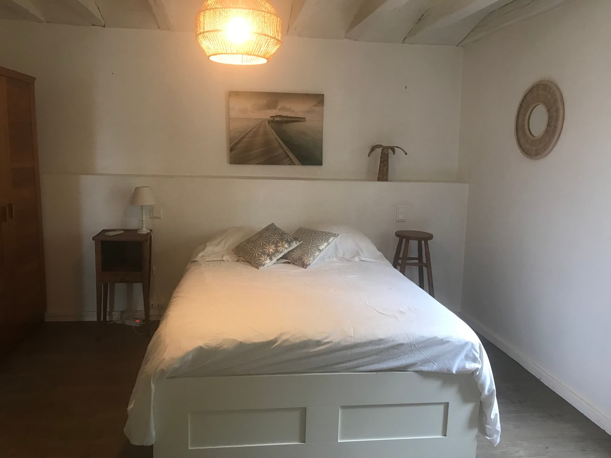 A spacious bed is positioned at the center of the room, adorned with two decorative pillows. A bedside lamp and a small table are located beside the bed. A wooden stool provides additional seating. Natural light filters in through a nearby window.