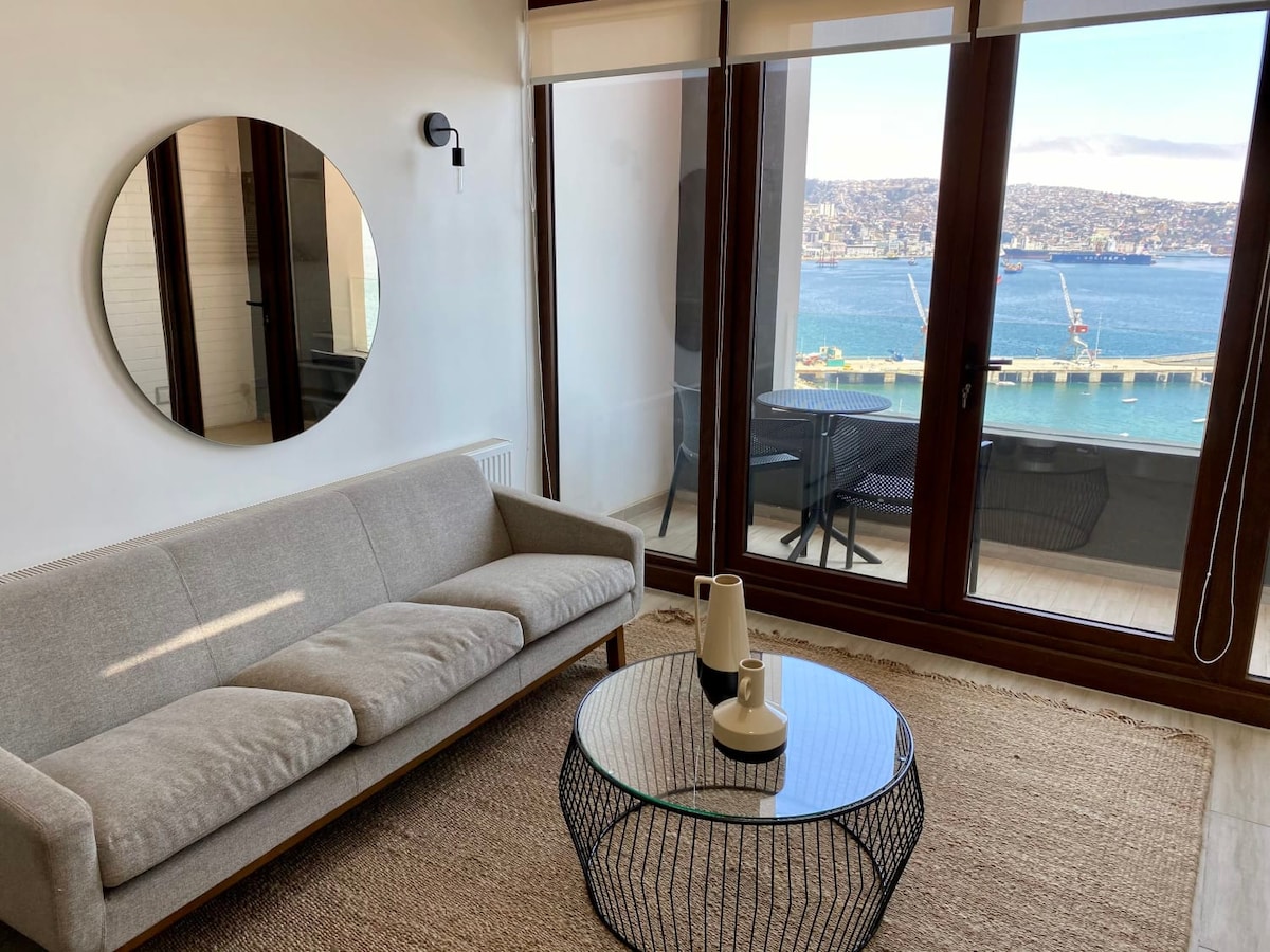 A modern living area features a light-colored sofa and a round glass coffee table, with a decorative vase on top. Large glass doors open to a balcony, offering ocean views. A circular mirror is mounted on the wall, enhancing the room's spacious feel.