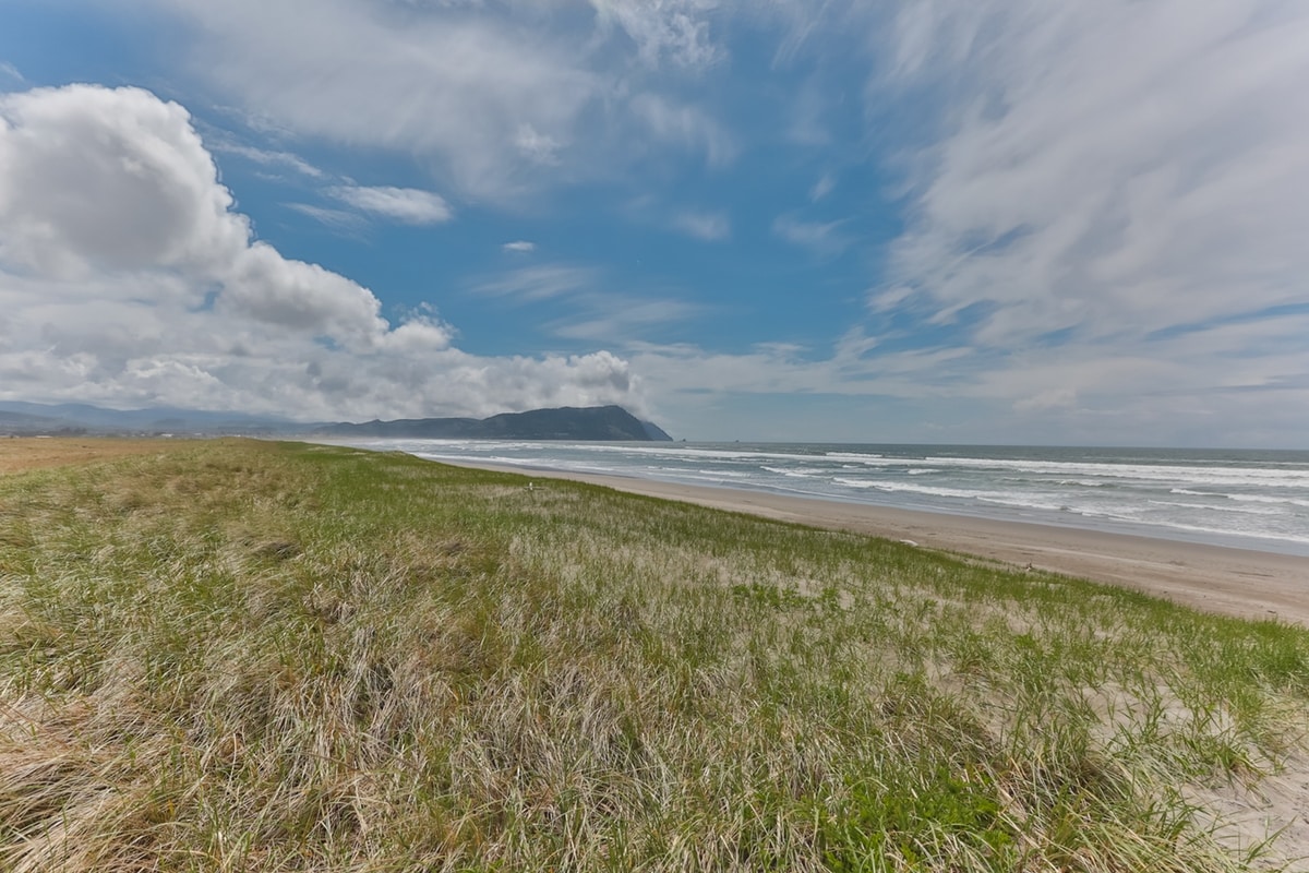 A wide sandy beach stretches under a blue sky, with soft waves lapping at the shore. Green grass and dunes line the beach, providing a natural border. The distant coastal mountains are faintly visible, adding depth to the serene landscape.