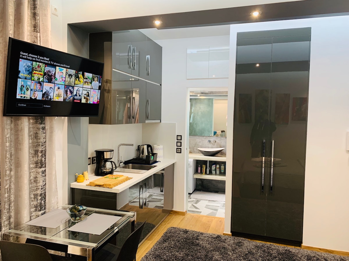 A modern kitchenette features sleek dark cabinetry, a compact dining area with a glass table, and an array of kitchen appliances including a kettle and coffee maker. A Smart TV is mounted on the wall, and a doorway leads to the bathroom area.