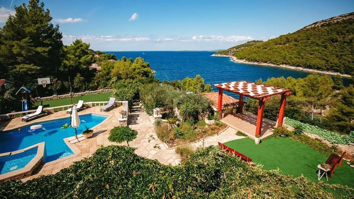 A scenic outdoor area features a swimming pool surrounded by sun loungers and lush greenery. A pergola offers shaded seating beside a well-kept garden. The blue sea and rolling hills provide a picturesque backdrop under a clear sky.