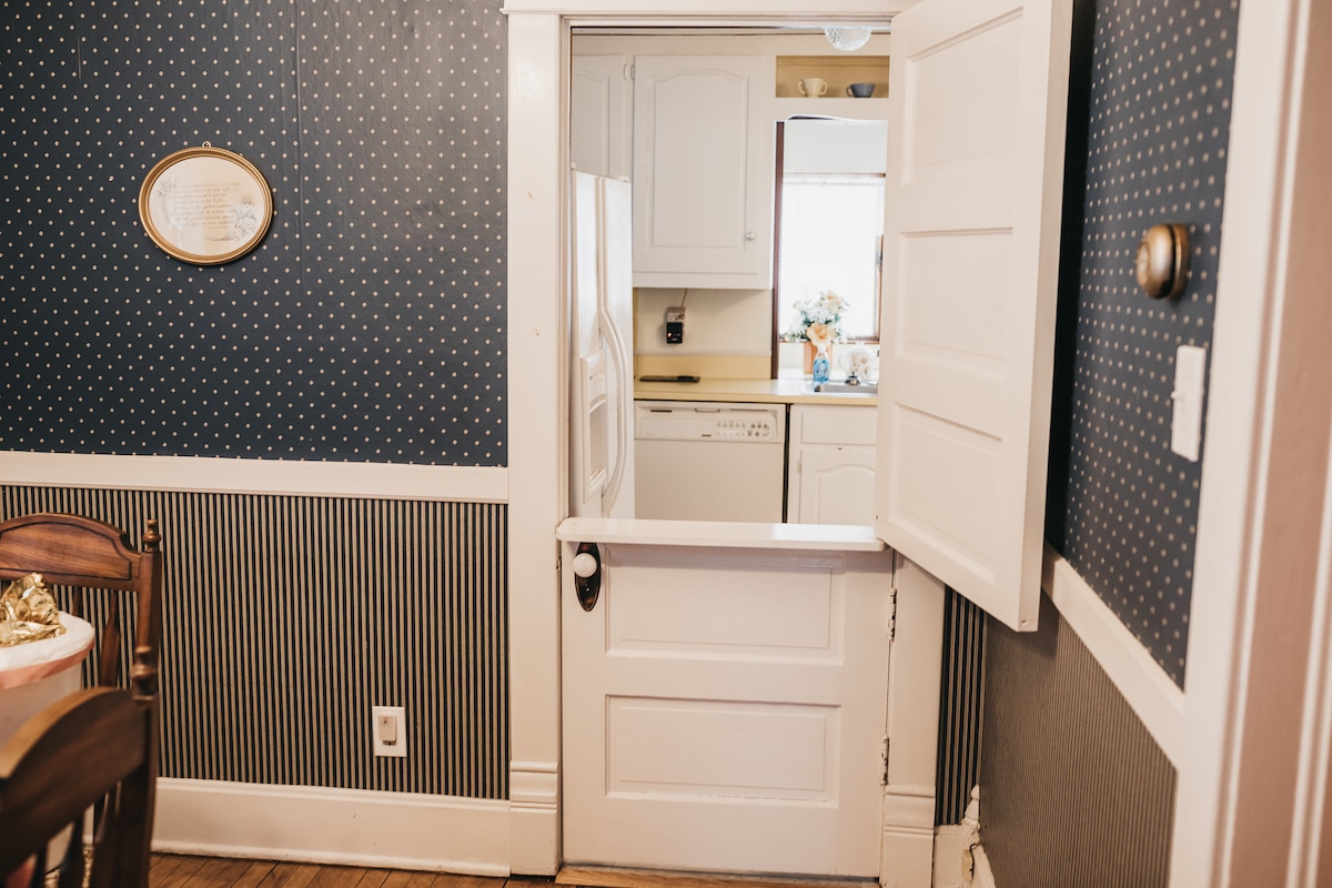 A charming entrance to a kitchen area is visible, featuring a classic door with a pass-through section. The wall is adorned with a polka dot pattern, complementing a decorative border. The white cabinetry and appliances are subtly seen, enhancing the welcoming atmosphere of the space.