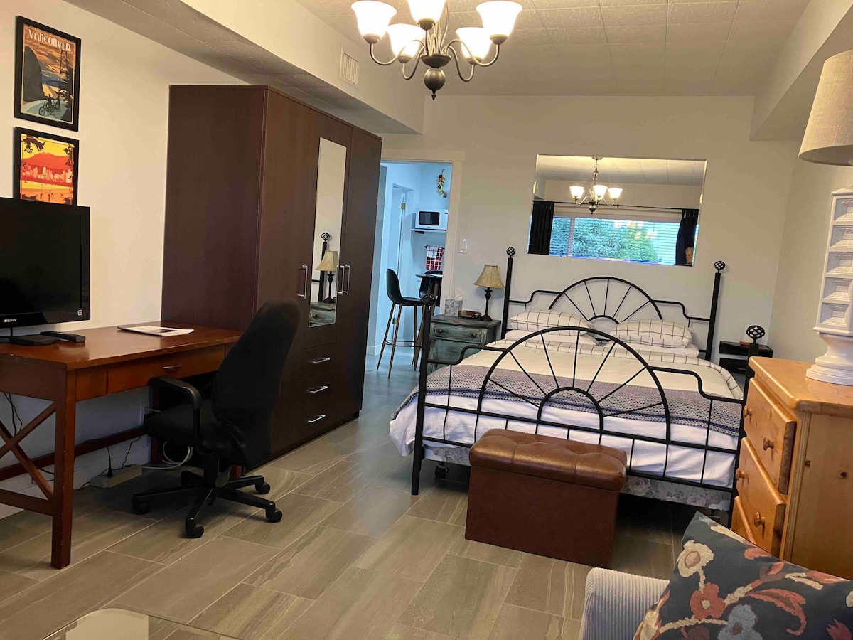 A functional living space includes a spacious bed with a decorative headboard and a cozy seating area. A desk and chair are positioned near a television, while a compact wardrobe and additional furniture provide storage. Natural light streams in from the window, enhancing the vibrant ambiance.