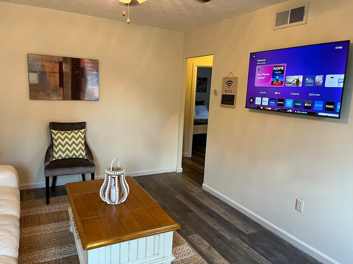 A cozy living area features a comfortable chair with a patterned cushion, a wooden coffee table, and a wall-mounted television displaying streaming options. Light-colored walls complement the textured area rug enhancing the space's inviting feel.