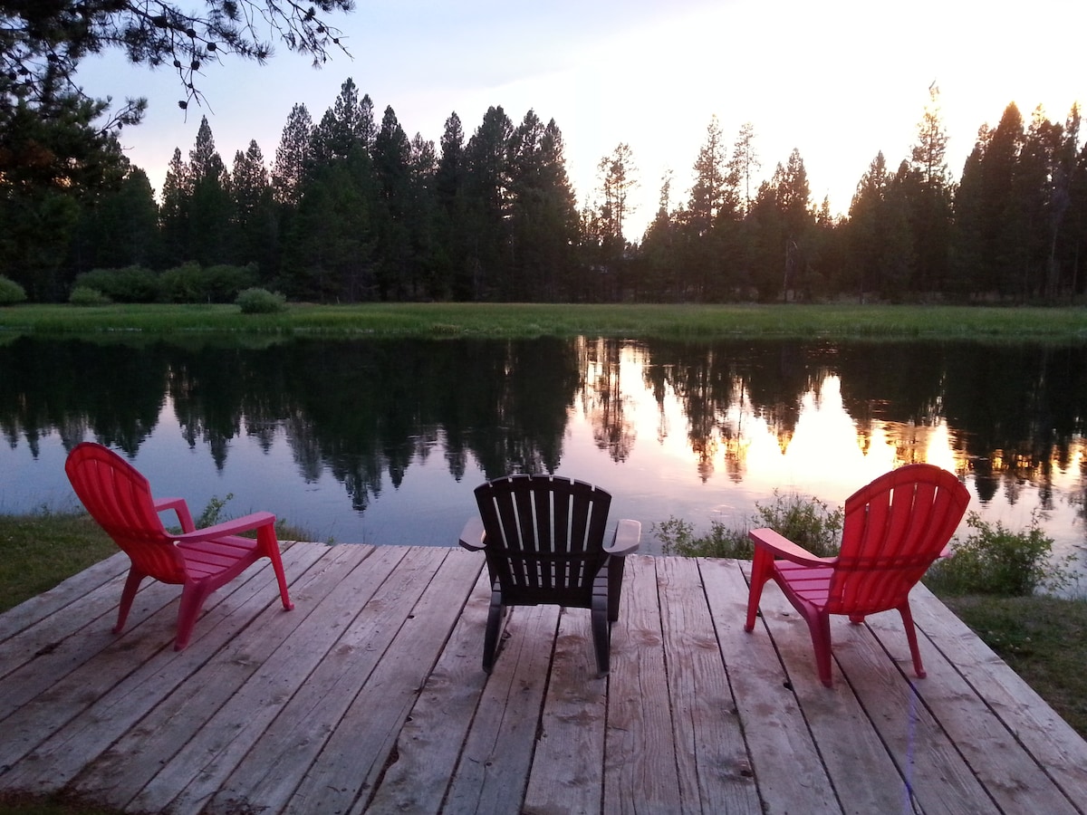 A wooden deck features three outdoor chairs, two in vibrant red and one in black, positioned to overlook a calm river. Surrounding greenery includes tall trees, with the sunset reflecting softly on the water's surface, creating a serene atmosphere.