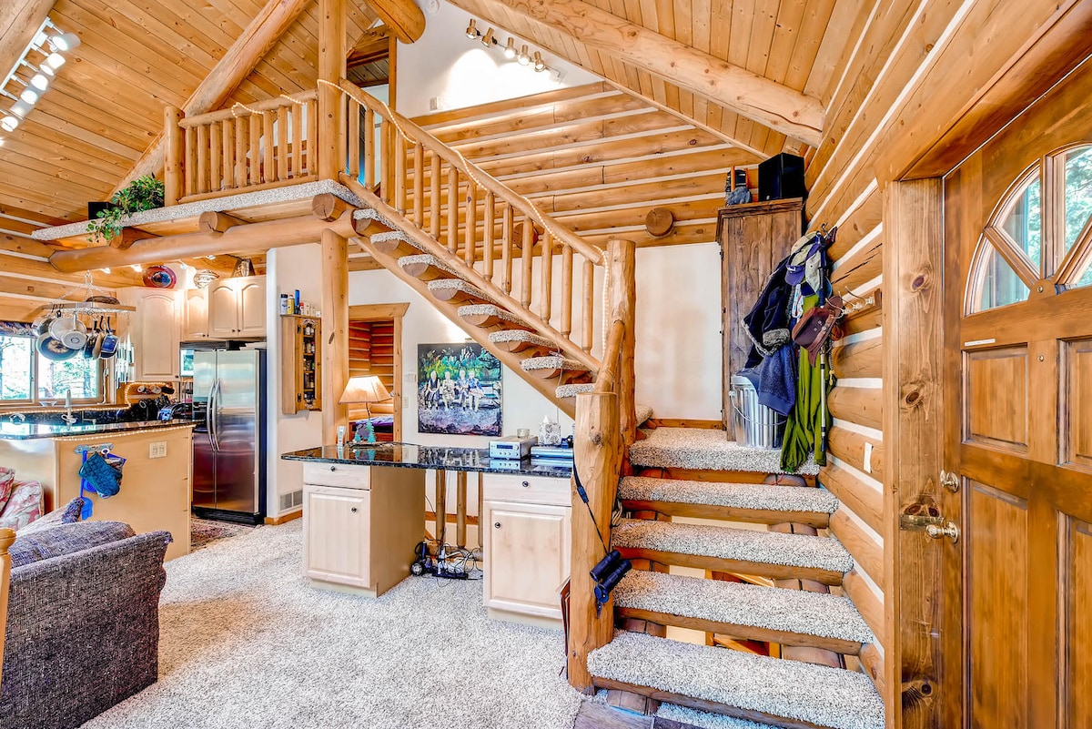 The interior features exposed wooden beams and a warm wooden staircase leading to the upper level. A spacious living area with a cozy couch and decorated with a wall-mounted TV creates a welcoming atmosphere. Natural light streams in through a large front door, highlighting the textured carpet.