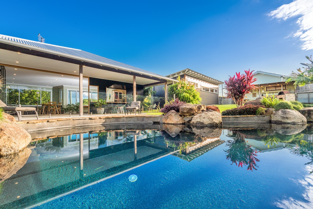 A tranquil freshwater rock pool reflects the surrounding greenery and sky. The sleek lines of the guesthouse can be seen through expansive glass walls, offering views of outdoor living spaces nestled among landscaped gardens. Vibrant plants add color along the poolside.