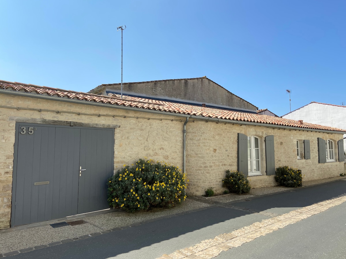 A charming stone facade of a house is displayed, featuring classic shutters and a gray garage door. Bright yellow flowers are positioned near the entrance, adding a touch of color. The clear blue sky complements the serene residential street.