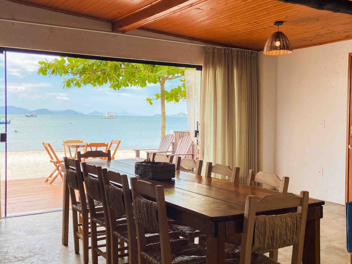 A spacious dining area features a long wooden table surrounded by woven chairs. Large glass doors provide a view of the tranquil beach and distant boats. Natural light filters through sheer curtains, complementing the warm tones of the wooden ceiling.