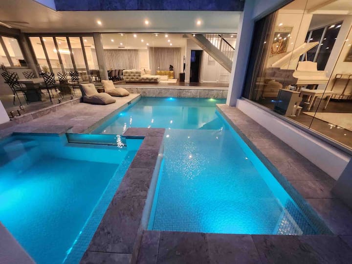 A modern indoor pool with glowing blue lights, surrounded by gray stone tiles. The pool area features cushioned lounge chairs, and large glass windows offer a view into a spacious, elegantly furnished living area with a piano.