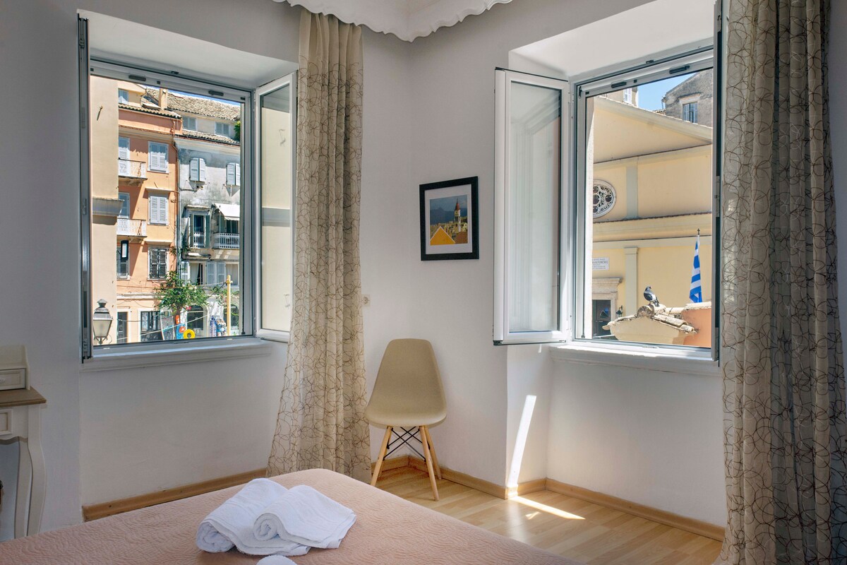 A bright bedroom is seen with dual windows offering views of the lively street outside. Light-colored curtains frame the windows, and a minimalist chair complements the space. A neatly arranged bed with fresh linens adds to the room's comfort.