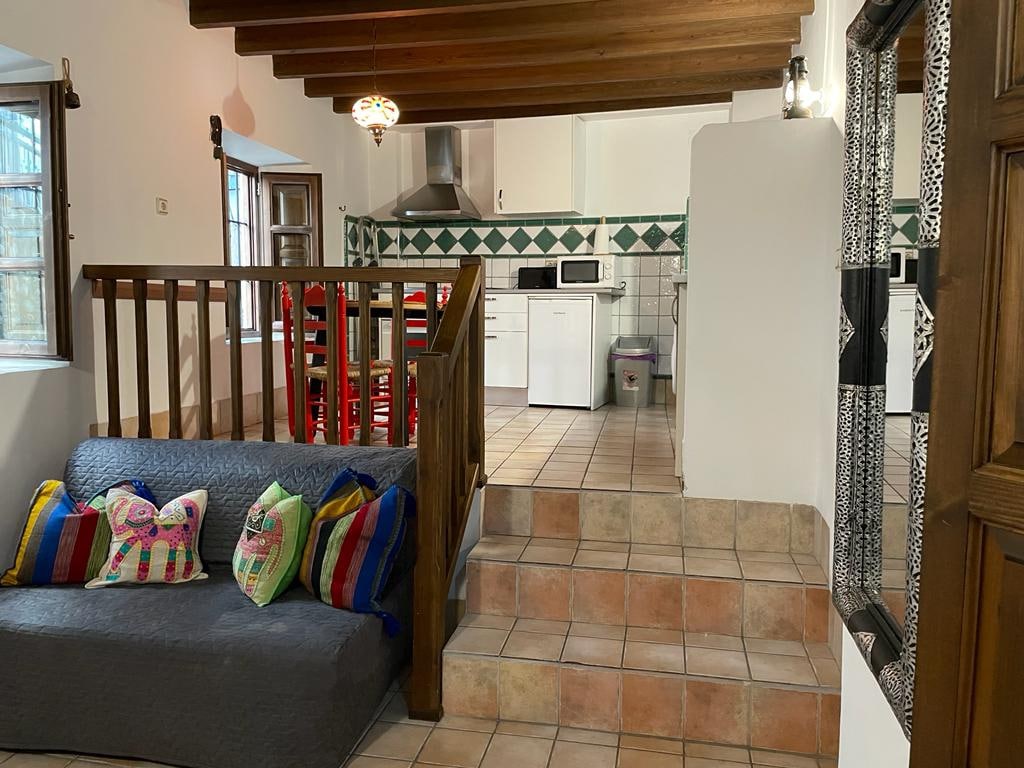 A cozy living area features a staircase leading to the kitchen, adorned with patterned tiles. A gray sofa is accompanied by vibrant, decorative pillows. A wooden railing complements the warm wooden beams overhead, while a table set with red chairs adds a pop of color.