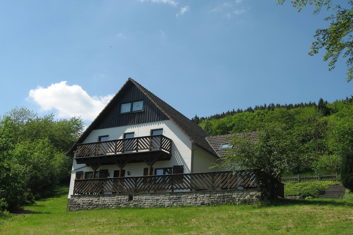 The exterior of the holiday home showcases a three-story structure, featuring a sloped roof and a balcony with wooden railings. Surrounded by greenery, the house is set against a clear blue sky, creating a serene outdoor environment.