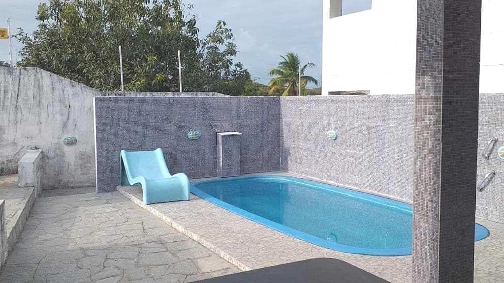 A rectangular swimming pool features clear blue water, surrounded by a mosaic tile wall. A light blue lounge chair is positioned beside the pool on a stone patio, while tropical vegetation is visible in the background, contributing to a relaxed outdoor setting.
