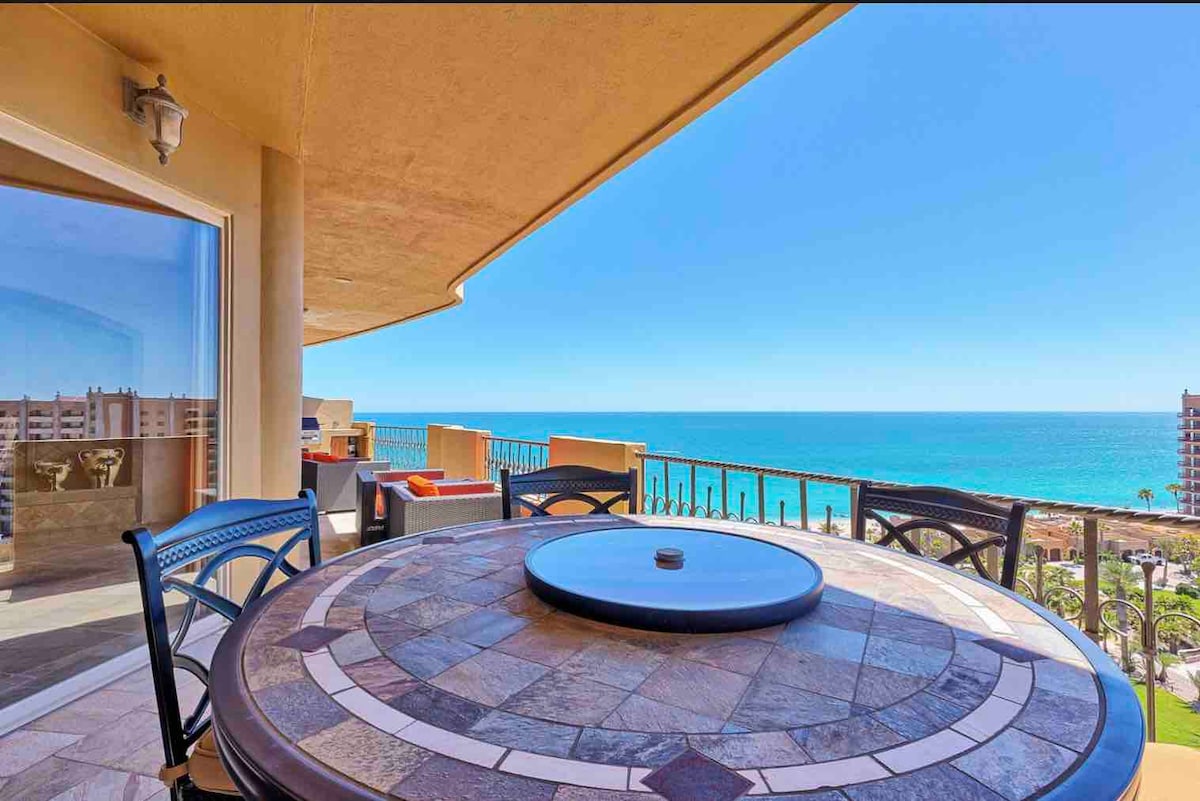 An outdoor dining table featuring a circular stone top sits on a balcony, offering expansive views of the ocean. The nearby railing complements the coastal scenery, while plush seating is arranged for an inviting atmosphere. Clear blue skies and calm waters enhance the tranquil setting.