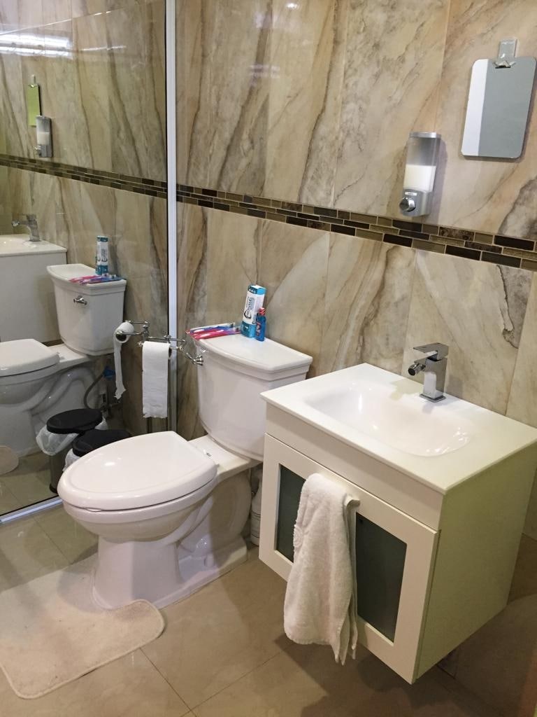 The bathroom features a modern design with marble-patterned walls and dark accent tiles. A white toilet and a sleek vanity with a sink are present, alongside a full-length mirror. Personal care items are neatly arranged, and a towel is hung on the vanity.