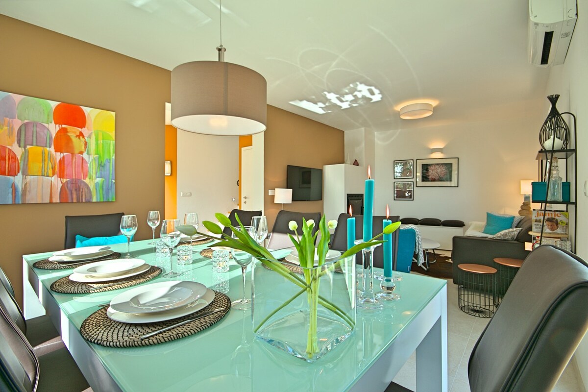 A dining area features a glass table set for eight, accented by tasteful tableware and a decorative vase with fresh flowers. The room is enhanced by vibrant wall art and a warm, inviting color palette, with a cozy living space visible in the background.