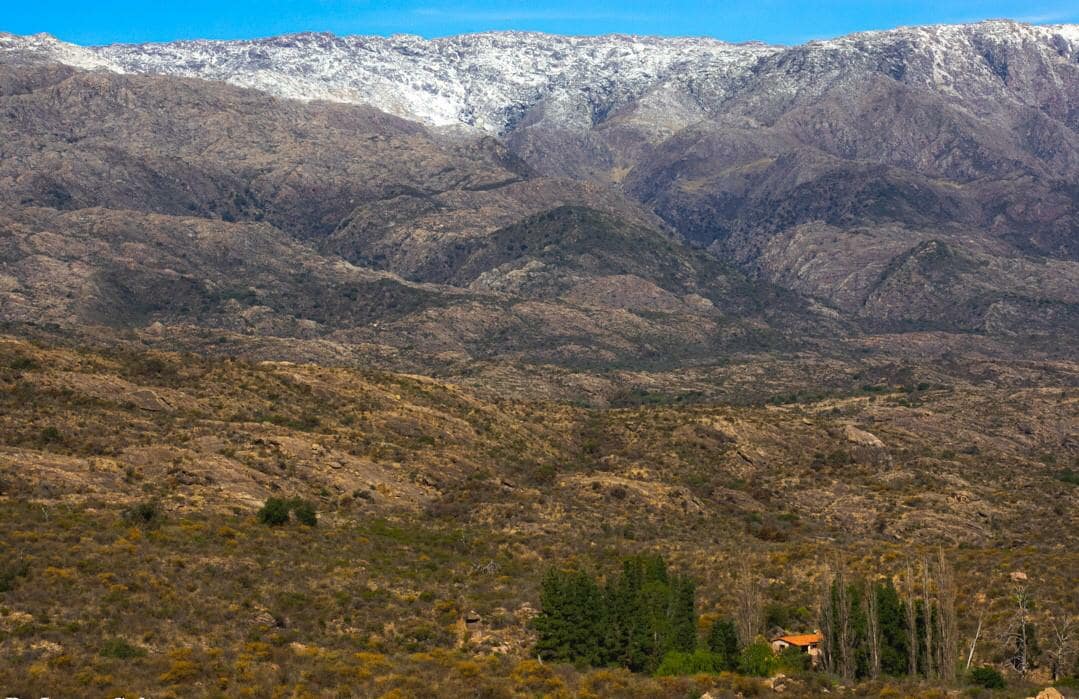 A wide expanse of rugged mountains is visible, with a snow-capped peak in the distance. The foreground features a gentle slope covered in dry grasses and shrubs, interspersed with clusters of trees. A small structure can be seen nestled among the vegetation.