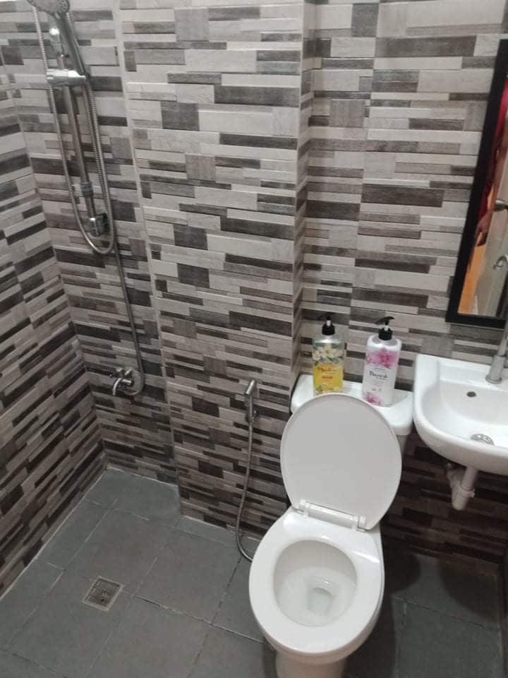 The modern bathroom features a shower with a sleek handheld fixture, surrounded by textured grey tile. A compact toilet is situated beside a small sink, with toiletries displayed on the countertop. The floor is tiled in a complementary grey, adding to the overall design.