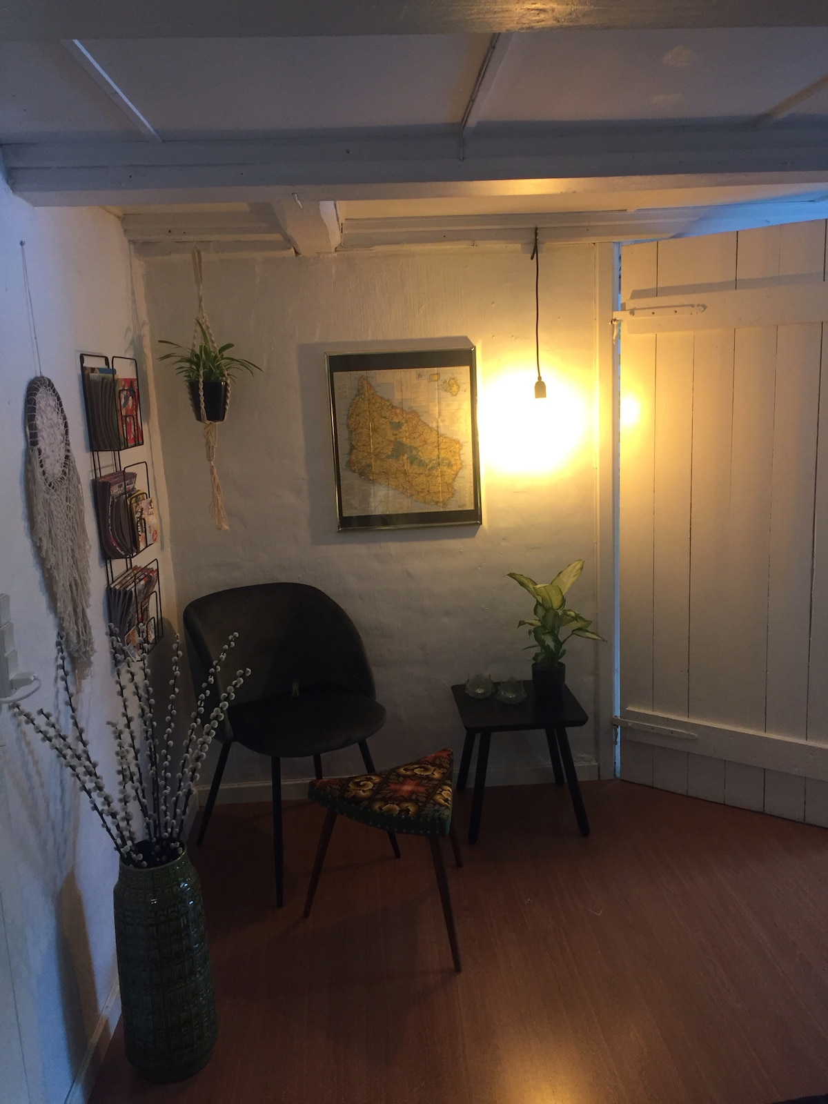 A cozy entryway features a black chair beside a small table adorned with a decorative plant. A vibrant, patterned stool adds color to the space. A framed map is hung on the wall, illuminated by a warm light, and a tall vase with branches completes the inviting setup.