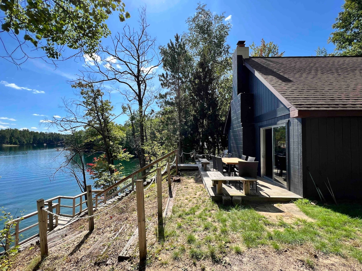 A spacious outdoor deck is located beside a serene lake, featuring a picnic table and additional seating. Lush greenery surrounds the area, with tall trees and a gentle slope leading down towards the water. The clear blue sky reflects off the calm lake surface.