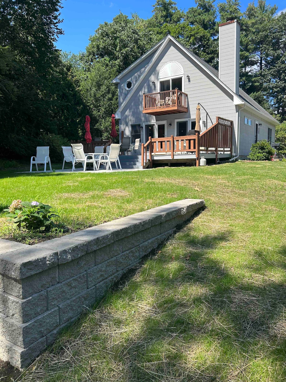 A two-story house is viewed from the yard, featuring a welcoming deck with wooden railings and seating. The lush green lawn is bordered by a stone wall, with bright red umbrellas overhead, offering shade. Tall trees provide a natural backdrop to the serene setting.