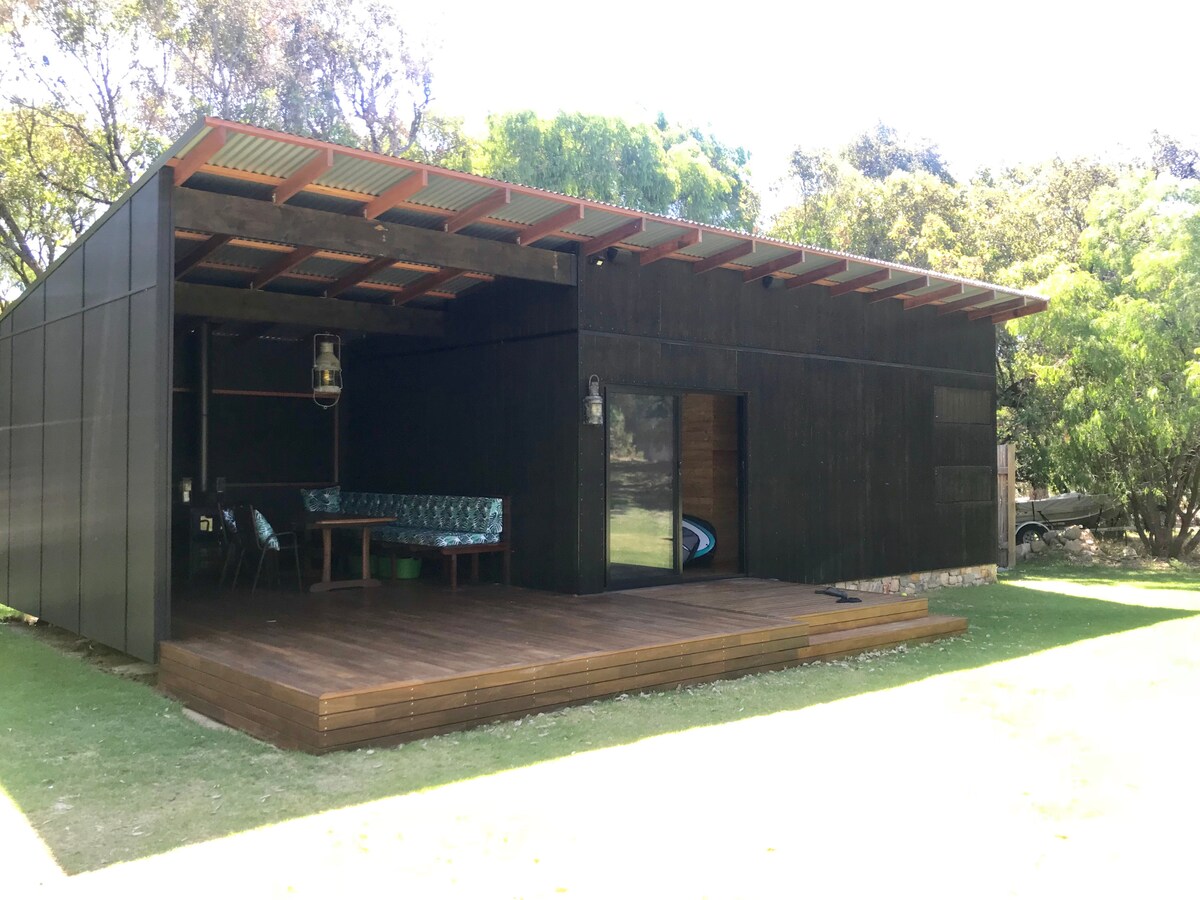 The exterior of the cabin showcases a modern design with a dark panel facade and a spacious wooden deck. A seating area with blue-patterned cushions is visible, sheltered by an overhang. Lush greenery surrounds the property, offering a tranquil setting.