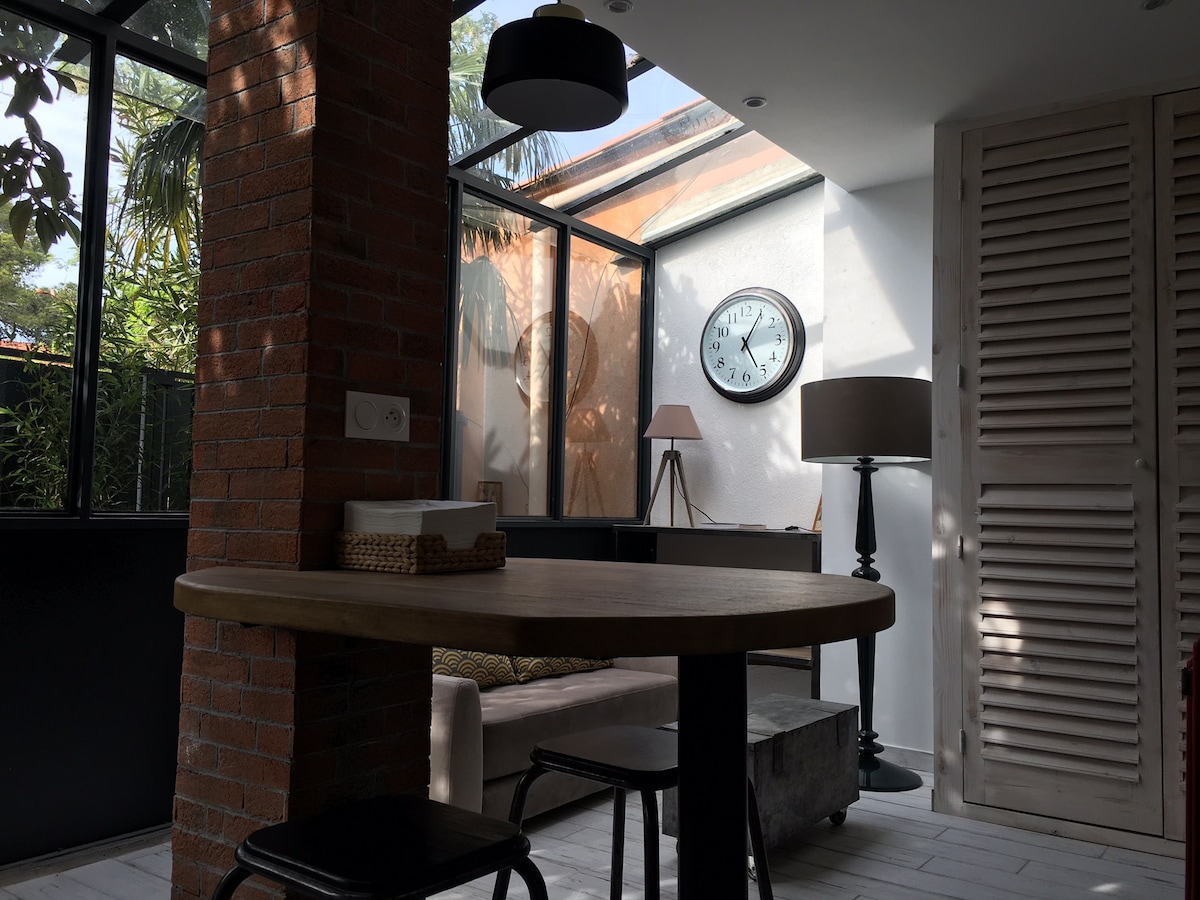A bright living area is featured with large glass walls, allowing natural light to fill the space. A round wooden table with black chairs is positioned in the foreground. A wall clock and a floor lamp are observed, adding functional elements to the room.