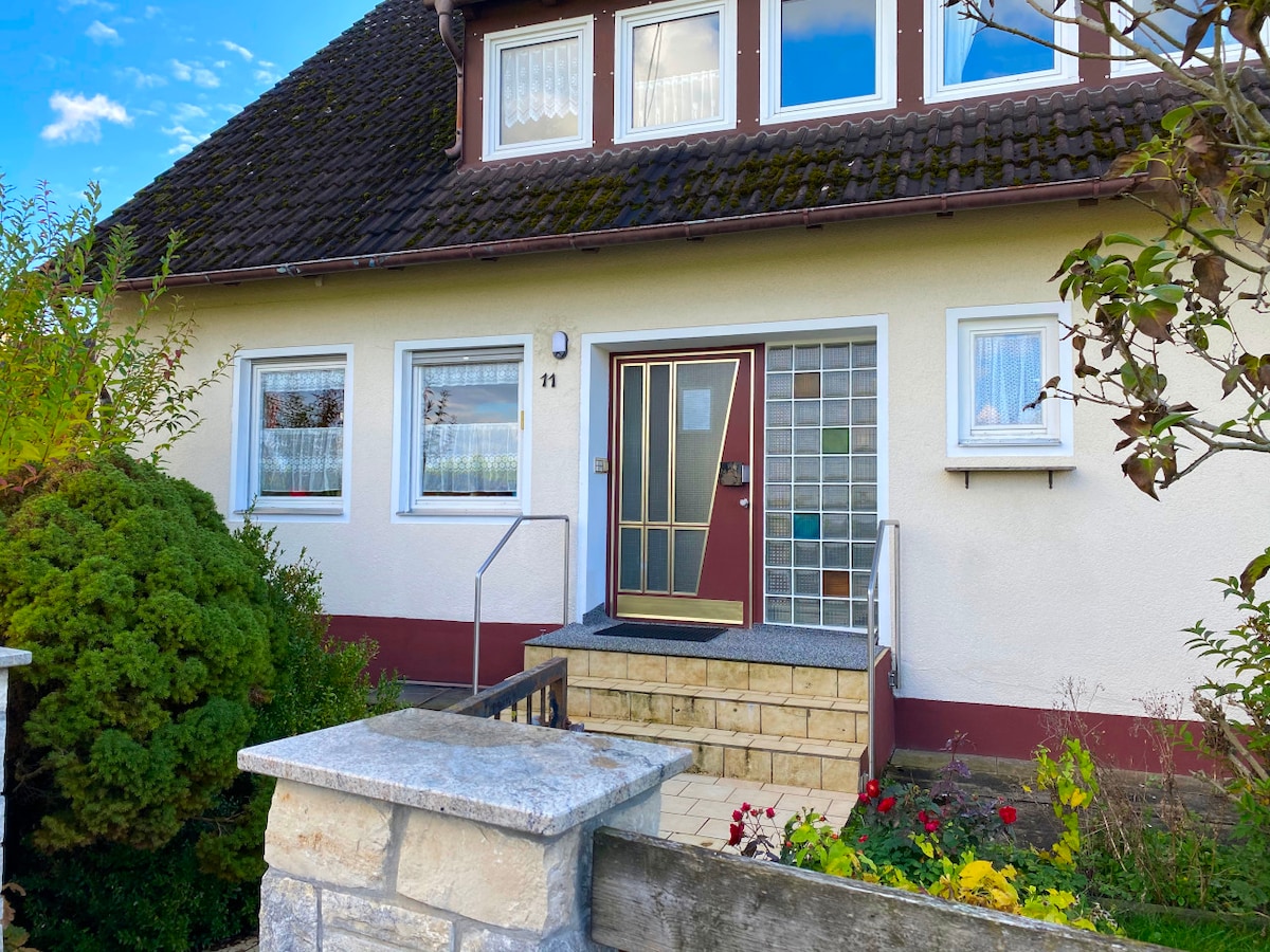A welcoming entrance is highlighted by a clean stone pathway leading to a front door with decorative glass panels. Lush greenery frames the entrance, adding a touch of nature, while colorful flowers are visible in the garden bed to the side.