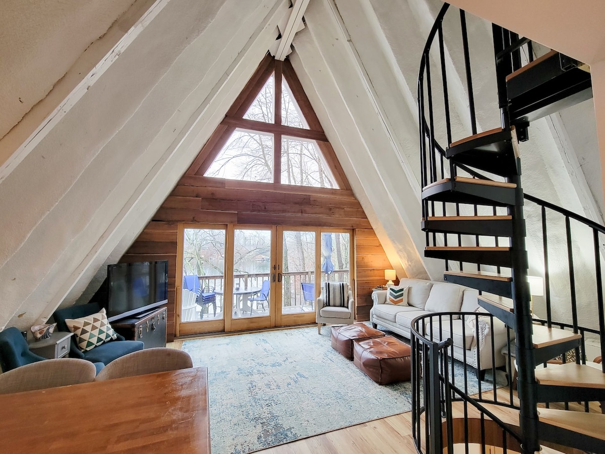 The spacious living area features an A-frame design with large triangular windows that flood the room with natural light. A comfortable sectional sofa and coffee table are arranged on a soft area rug, while a dining table is visible in the foreground. A spiral staircase leads to the loft above.