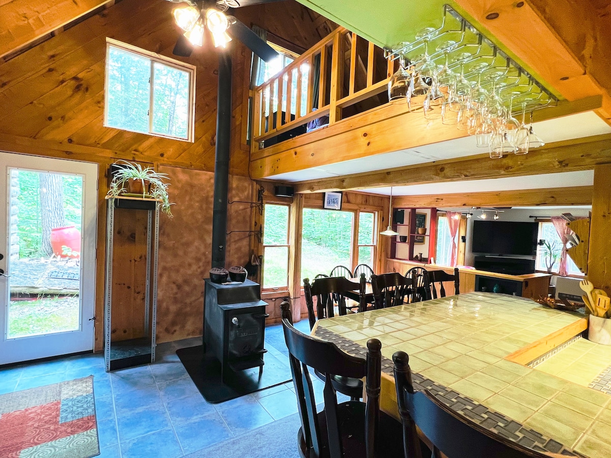 An open dining area with a large tiled breakfast bar and dark wooden chairs invites communal meals. A stylish wood stove adds warmth, while natural light streams through multiple windows. The vaulted ceiling enhances the spacious feel, highlighted by rustic wooden beams.