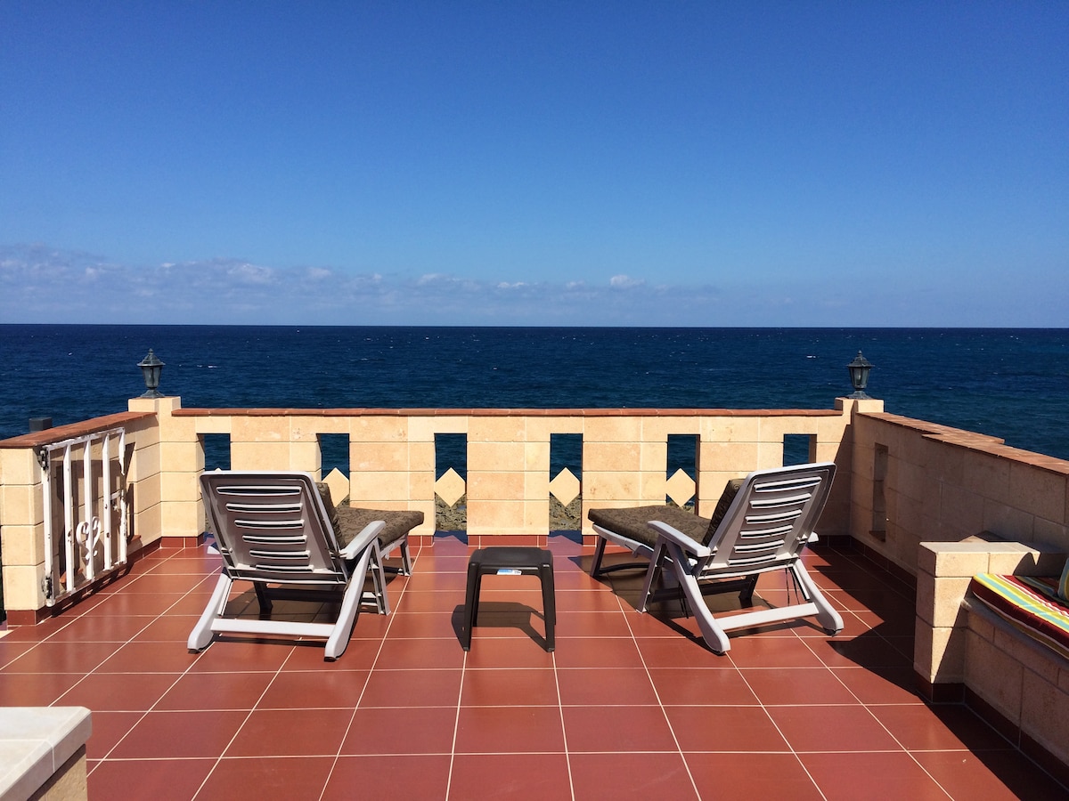 Two loungers are positioned on a tiled terrace, overlooking the Caribbean Sea. The surrounding area features a low stone wall, and the tranquil blue waters extend to the horizon, under a clear sky with a few scattered clouds.
