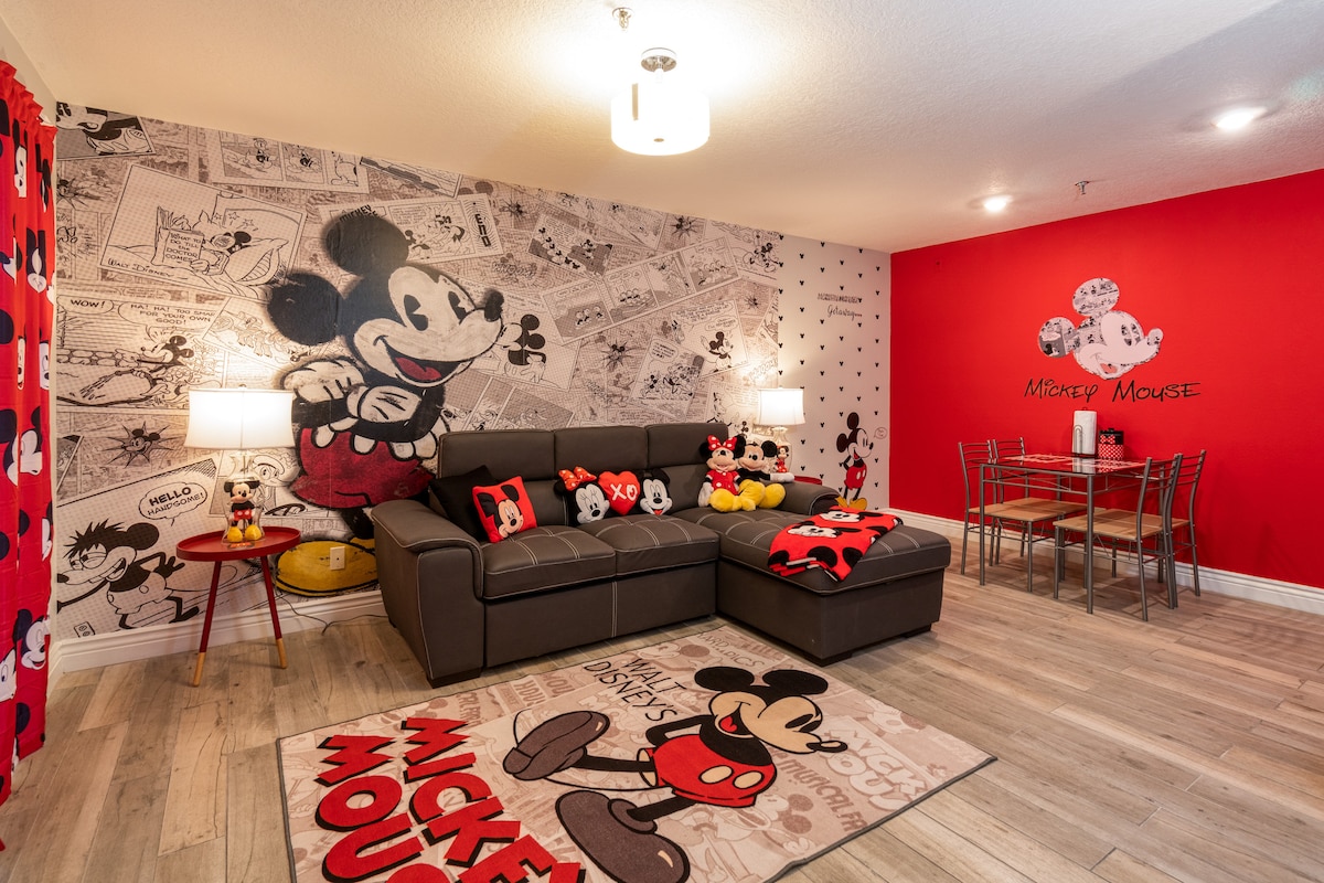 A modern living area features a large gray sectional sofa adorned with Mickey Mouse themed pillows. A playful rug with Mickey Mouse designs rests on the light wooden floor. Bright red walls are complemented by wall art showcasing iconic Mickey imagery, while a glass dining table enhances the space.