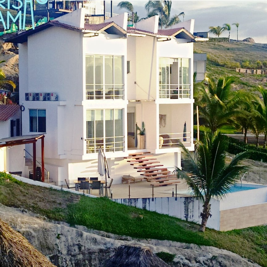 A modern, multi-level house features sleek glass windows and a spacious terrace with lounge chairs. A private infinity pool is visible, complemented by lush greenery and palm trees. The building's architecture reflects a minimalist design, enhancing the serene coastal surroundings.